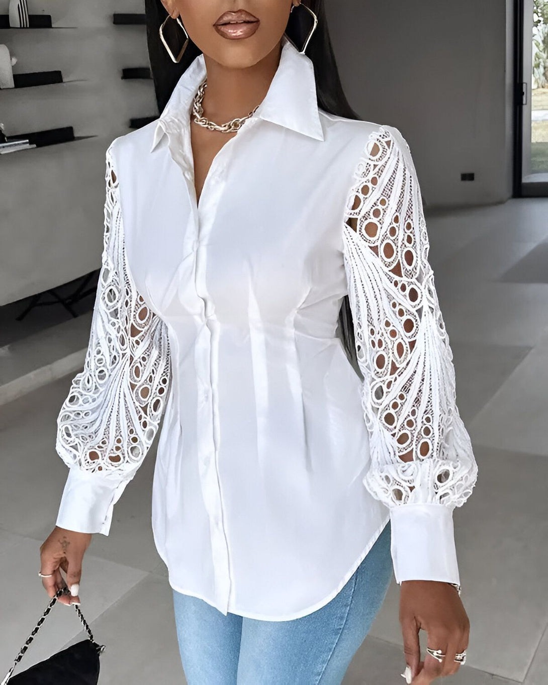 Une femme porte une chemise blanche avec des manches en dentelle. Elle est debout dans un bureau, et porte un jean bleu et des talons noirs.
