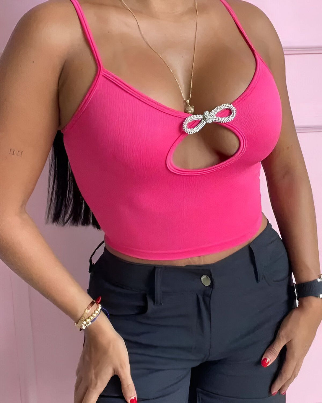 Une femme porte un haut rose avec un nœud en strass au niveau de la poitrine. Elle porte également un pantalon noir et des talons noirs.