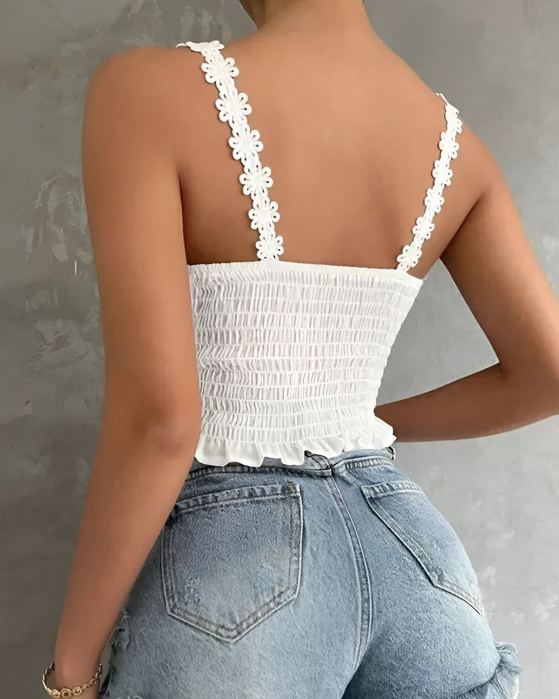 Une jeune femme porte un haut blanc à bretelles fines et un jean bleu taille haute. Le haut est froncé sur le devant et possède des détails en forme de fleurs sur les bretelles.