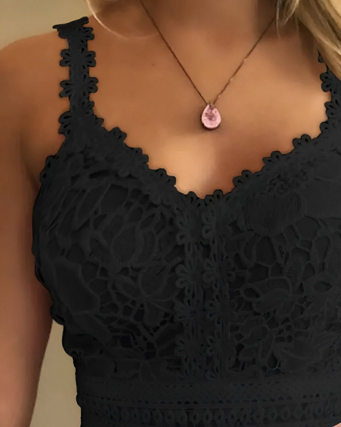 Une femme porte un haut en dentelle noir. Le haut est court et a un décolleté en V. La femme porte également un collier avec un pendentif en pierre rose.