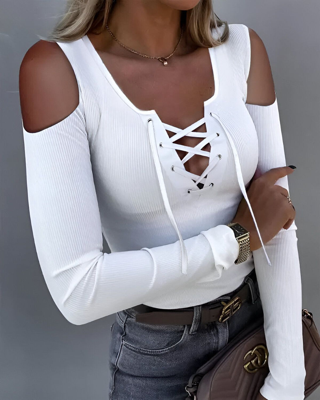 Une femme porte un haut blanc à manches longues avec un laçage au niveau du décolleté. Elle porte également un jean gris et une ceinture marron.