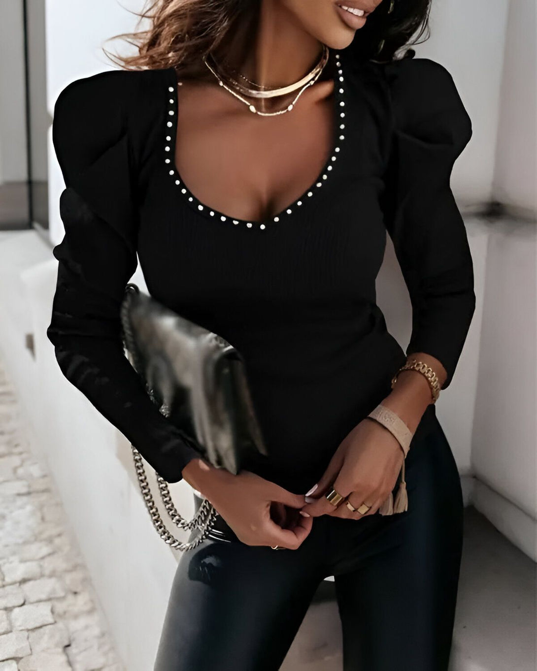 Une femme porte un haut noir à manches bouffantes et un jean noir. Elle a les cheveux longs et bruns et porte un collier. Elle est debout dans la rue et sourit.