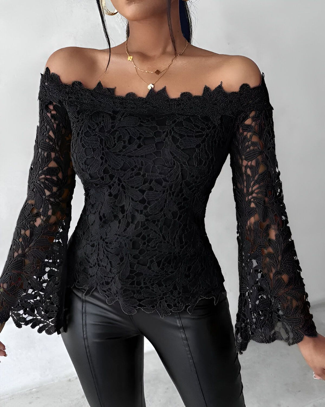 Une femme porte un haut en dentelle noir à épaules dénudées. Le haut est cintré à la taille et possède des manches longues évasées. Elle porte également un pantalon en cuir noir.