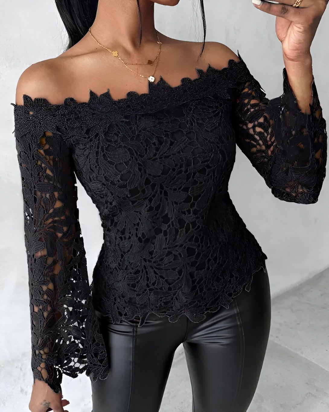 Une femme porte un chemisier noir en dentelle à épaules dénudées. Le chemisier est transparent et laisse entrevoir sa peau. Elle porte également un pantalon en cuir noir.