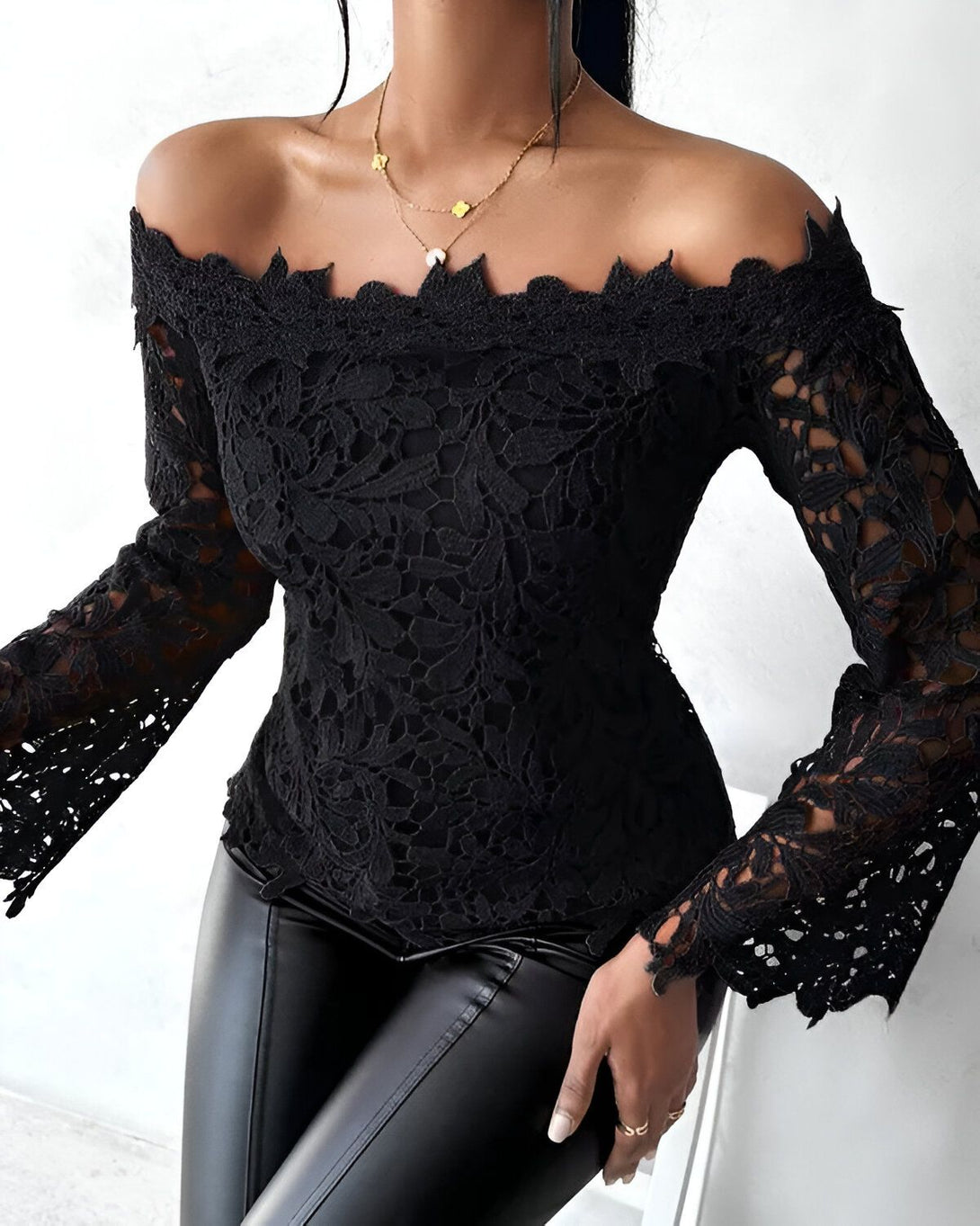 Une femme porte un chemisier noir en dentelle à épaules dénudées. Le chemisier est ajusté et a des manches longues. Elle porte également un pantalon en cuir noir.