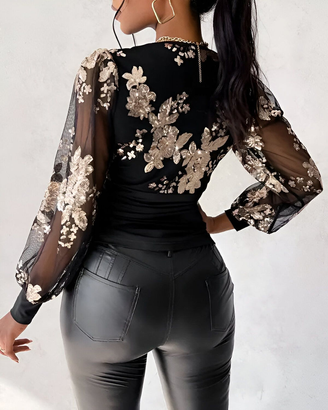 Une femme porte un chemisier noir à manches longues en résille brodées de sequins dorés et un pantalon en cuir noir.