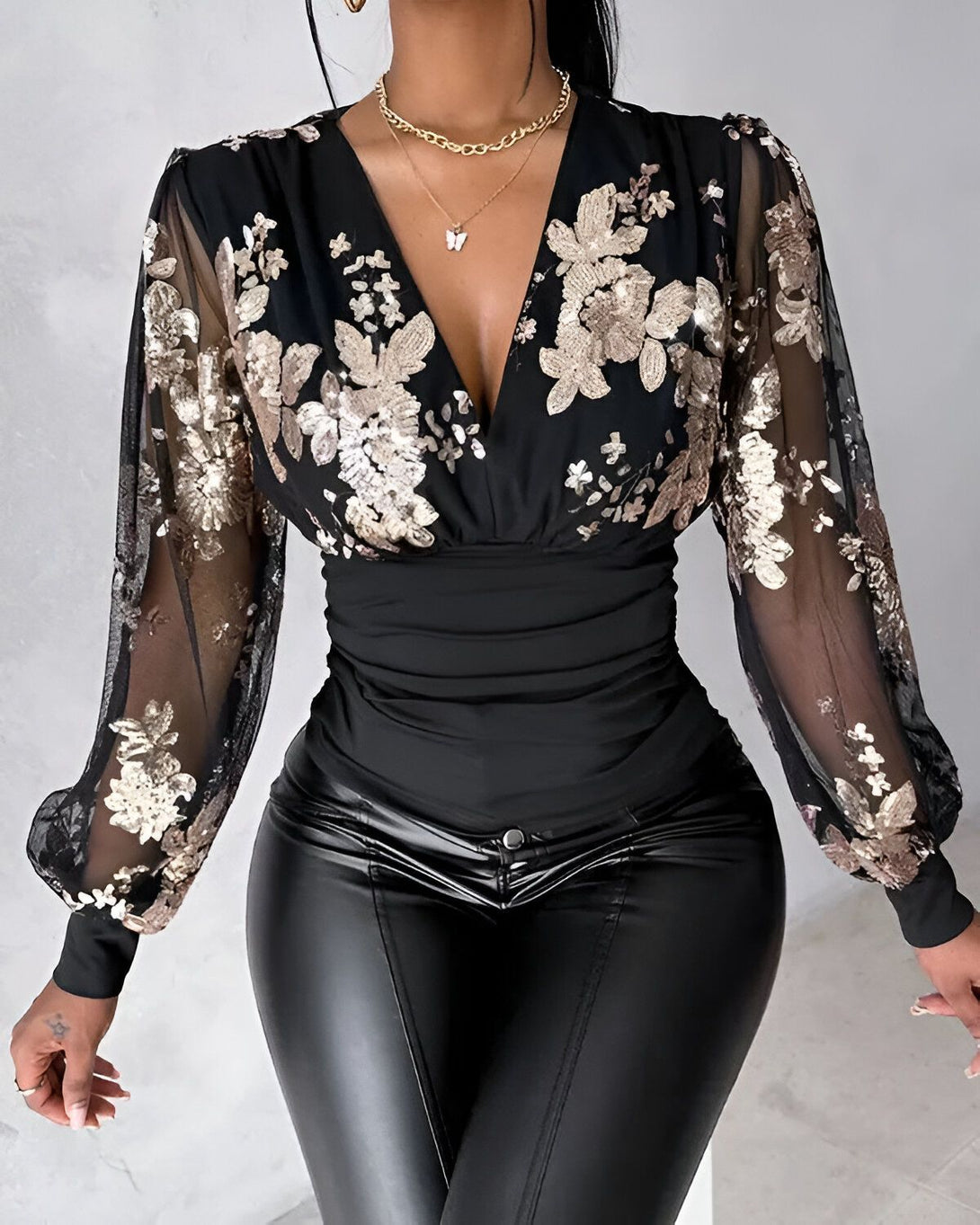 Une femme porte un chemisier noir à manches longues et un pantalon en cuir noir. Le chemisier est en mesh transparent avec des fleurs brodées de sequins dorés.
