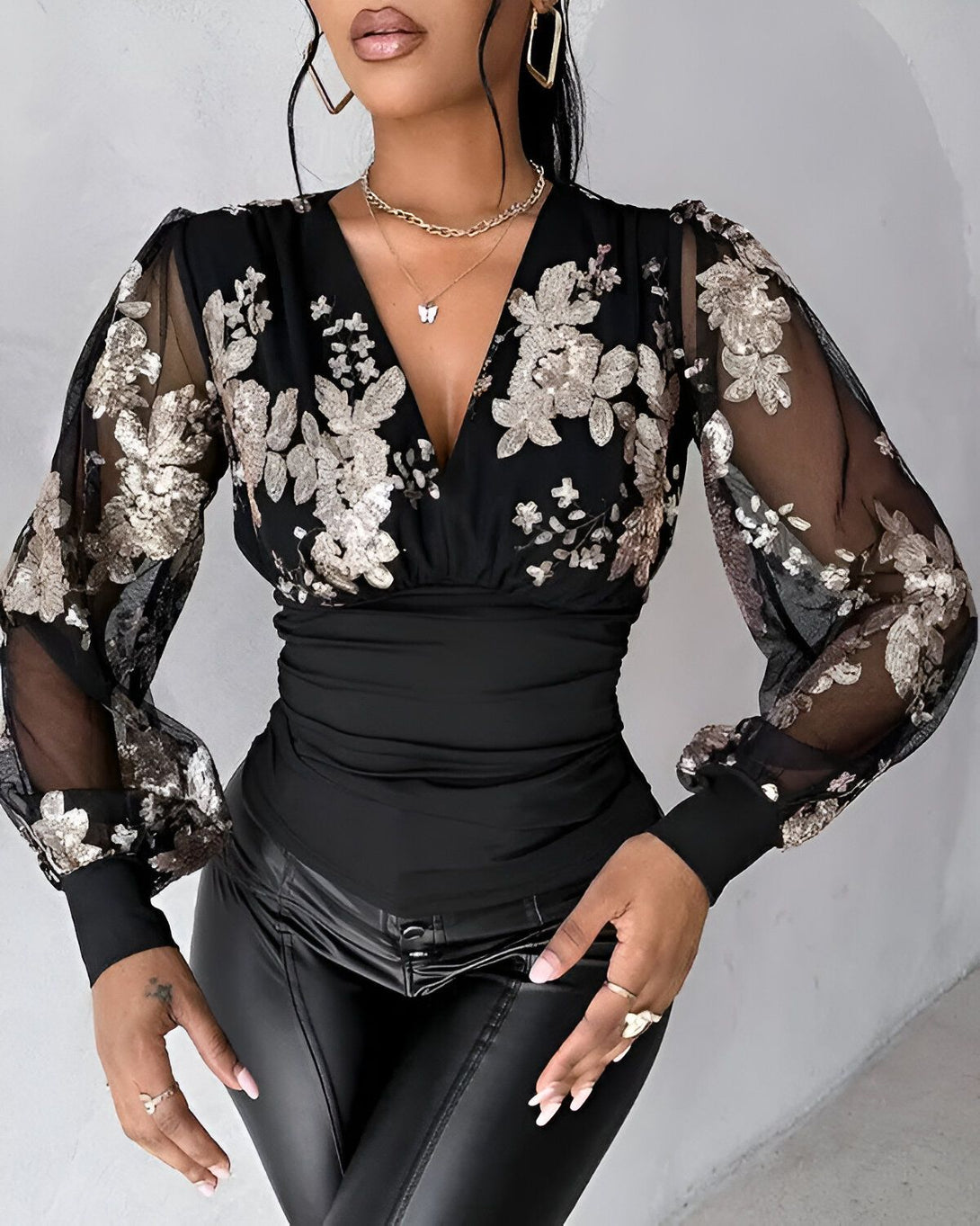 Une femme porte un chemisier noir à manches longues et transparentes. Le chemisier est orné de fleurs dorées et de sequins. La femme porte également un pantalon en cuir noir et un collier doré.