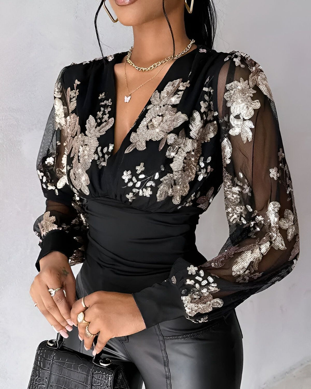 Une femme porte un chemisier noir à manches longues et transparentes. Le chemisier est orné de sequins dorés en forme de fleurs. La femme porte également un pantalon en cuir noir et des talons noirs.