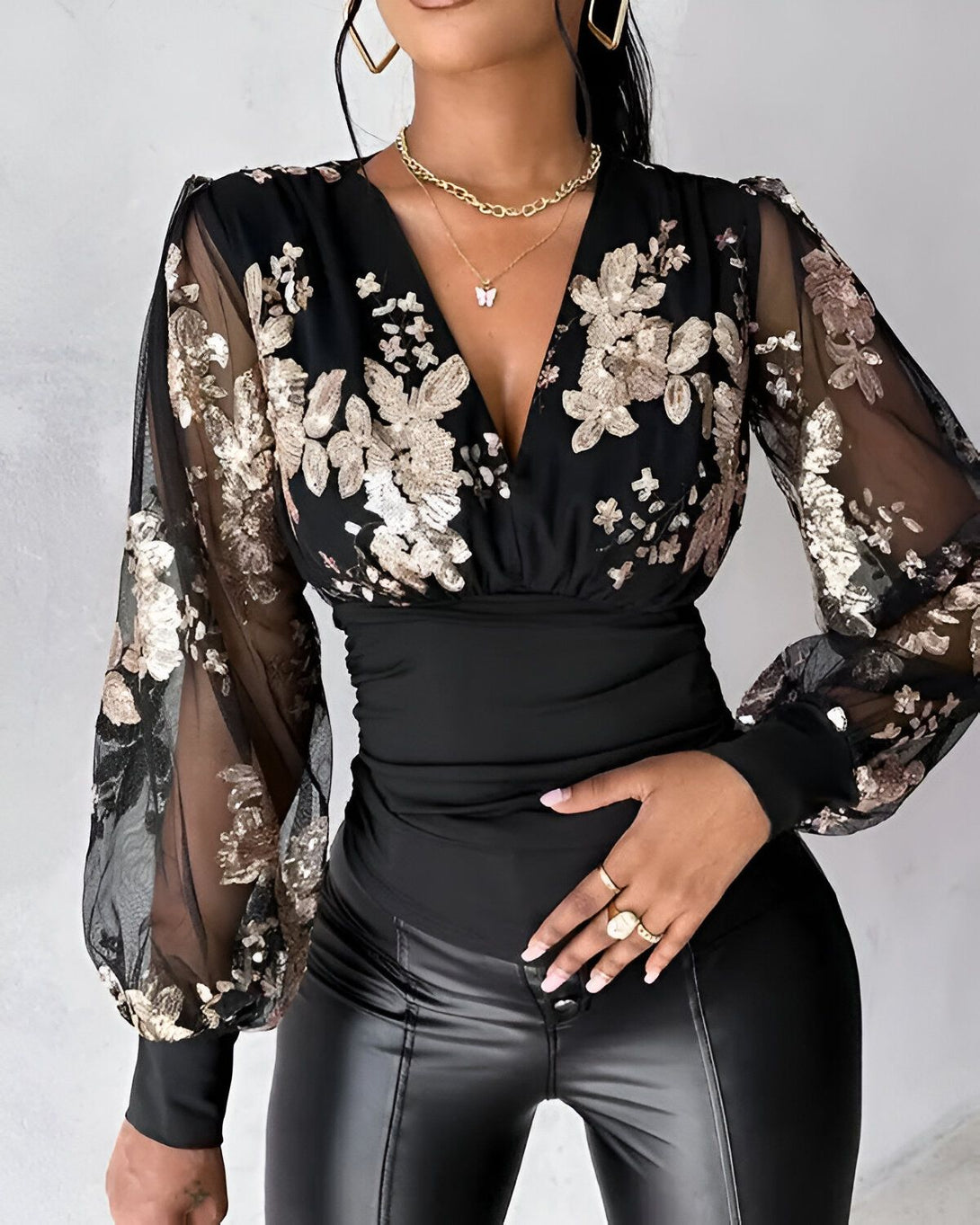 Une femme porte un chemisier noir à manches longues et transparentes. Le chemisier est orné de sequins dorés et de fleurs brodées. Elle porte également un pantalon en cuir noir et un collier doré.