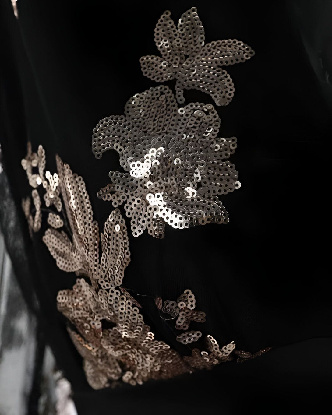 Une image en gros plan de sequins argentés cousus sur un tissu noir. Les sequins sont cousus en forme de fleurs et de feuilles.