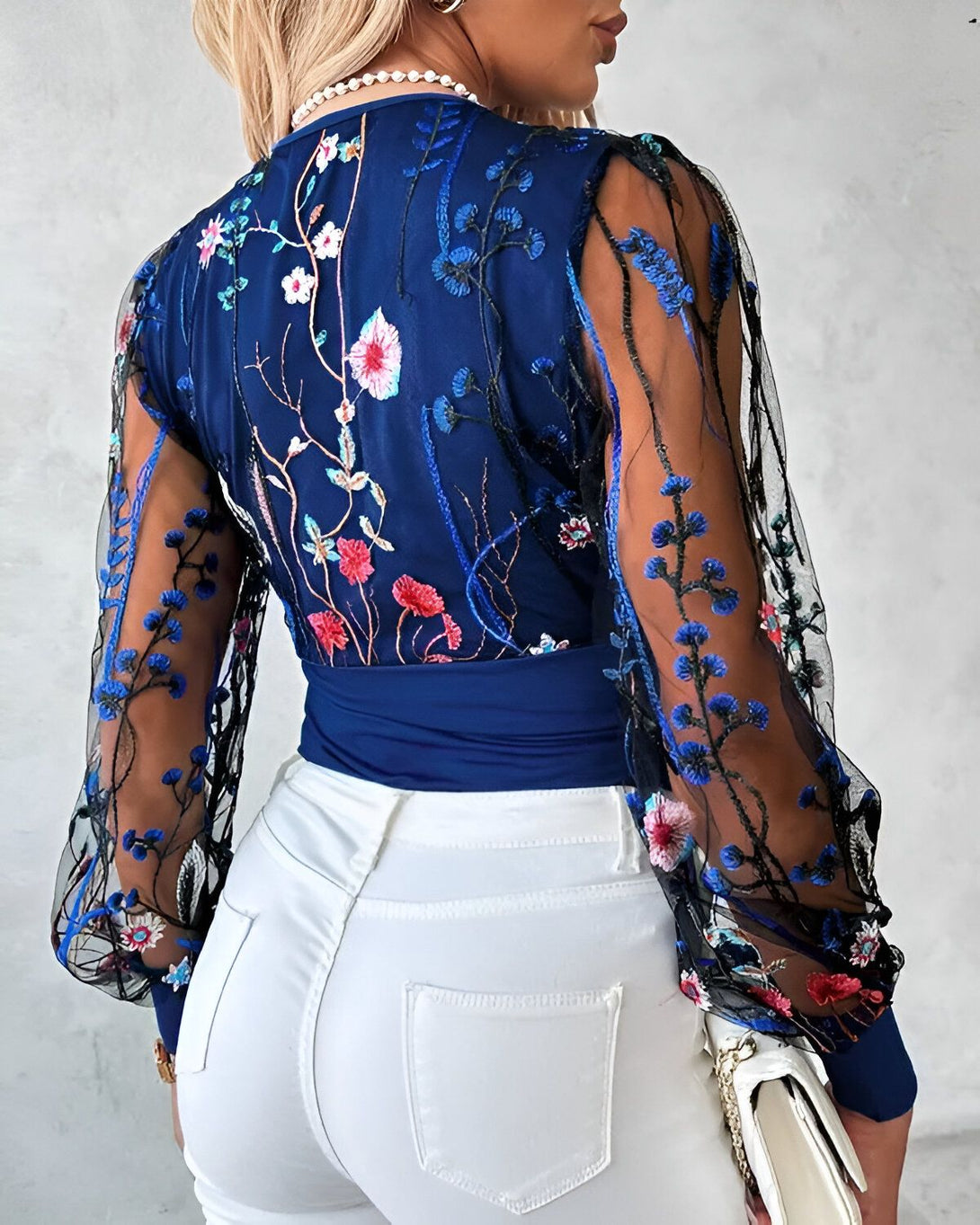 Une femme porte un chemisier bleu avec des fleurs brodées et des manches en résille noire. Elle porte également un jean blanc et des talons noirs.