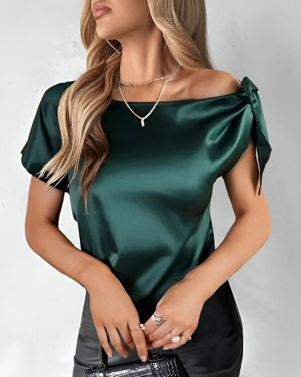 Une jeune femme porte un chemisier en satin vert. Le chemisier a des manches courtes et un col bateau. La femme a les cheveux longs et blonds. Elle porte également un collier et des boucles d'oreilles.
