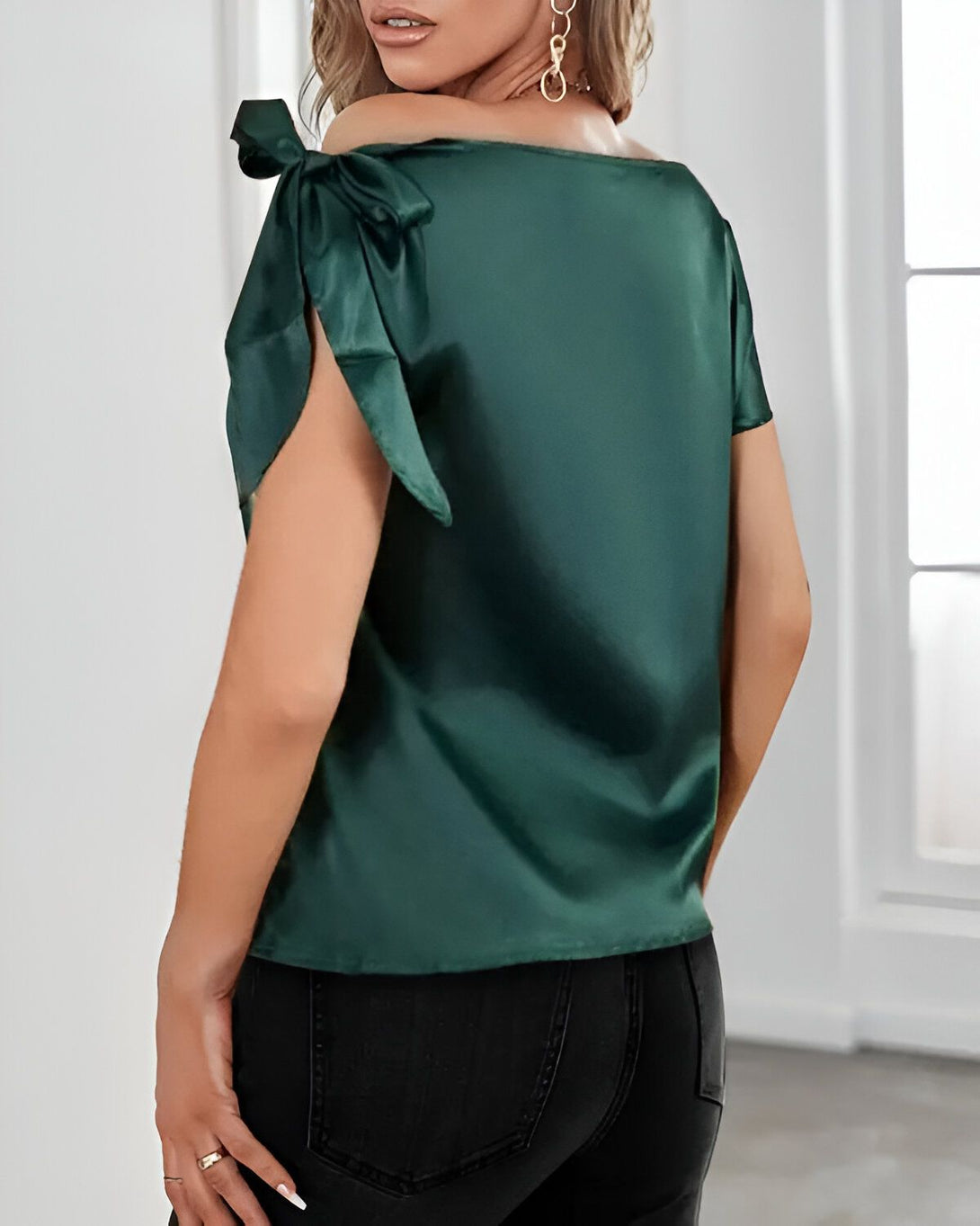 Une femme porte un chemisier vert en satin avec des nœuds aux épaules. Le chemisier est rentré dans un jean noir.