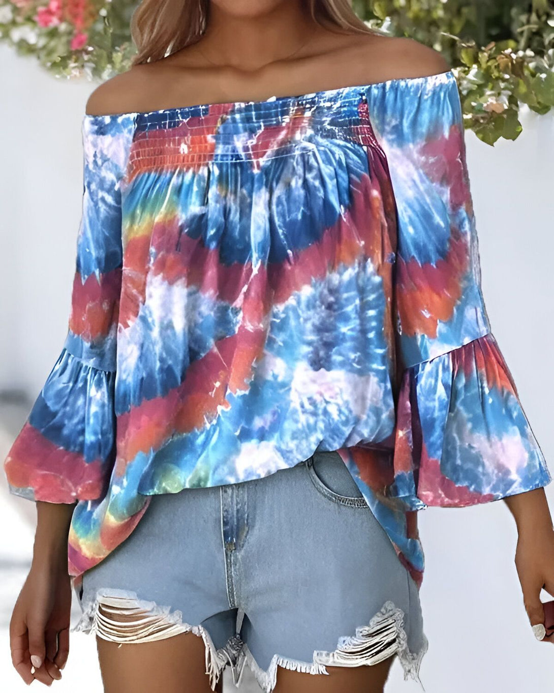 Une femme porte un chemisier tie-dye aux épaules dénudées et des shorts en jean déchirés. Le chemisier est bleu, rose et orange, et les shorts sont bleu clair. La femme a les cheveux longs et blonds et elle est souriante.