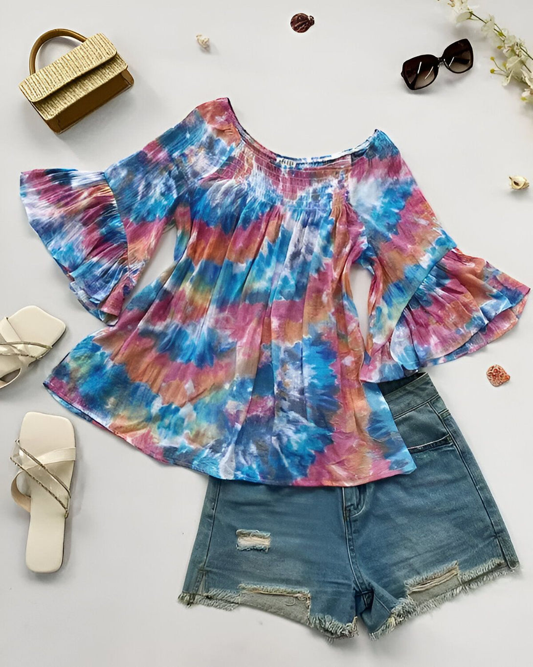 Un chemisier tie-dye rose, bleu et orange avec des manches bouffantes et un short en jean bleu déchiré. À côté se trouve une paire de sandales blanches et un sac à main marron.