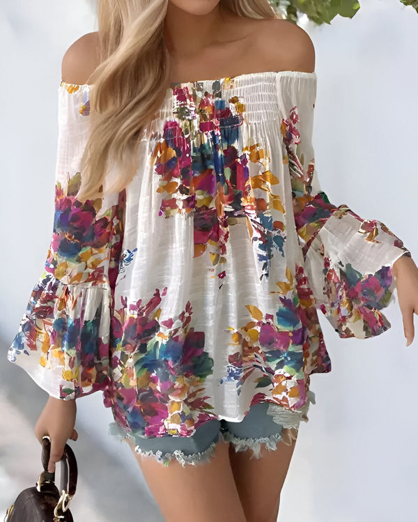 Une femme porte un chemisier blanc à épaules dénudées avec des fleurs colorées. Le chemisier est ample et a des manches bouffantes. La femme est blonde et porte un short en jean bleu. Elle porte également un sac à main marron.