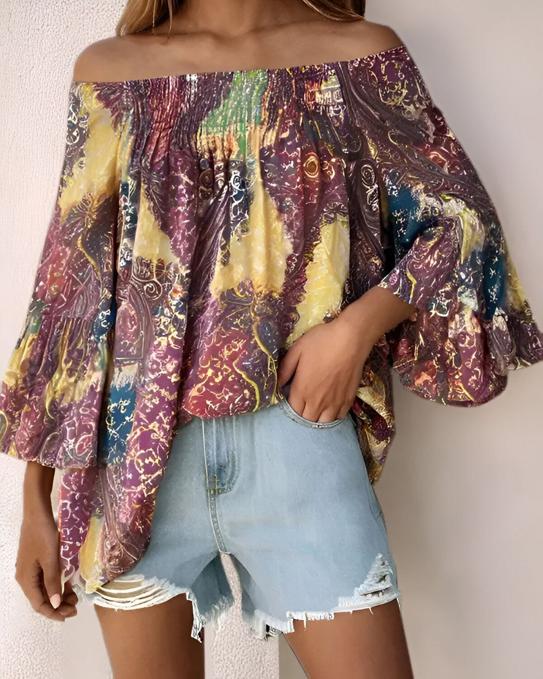Une femme porte un chemisier jaune et violet à épaules dénudées et des manches bouffantes. Elle porte également un short en jean bleu clair déchiré.