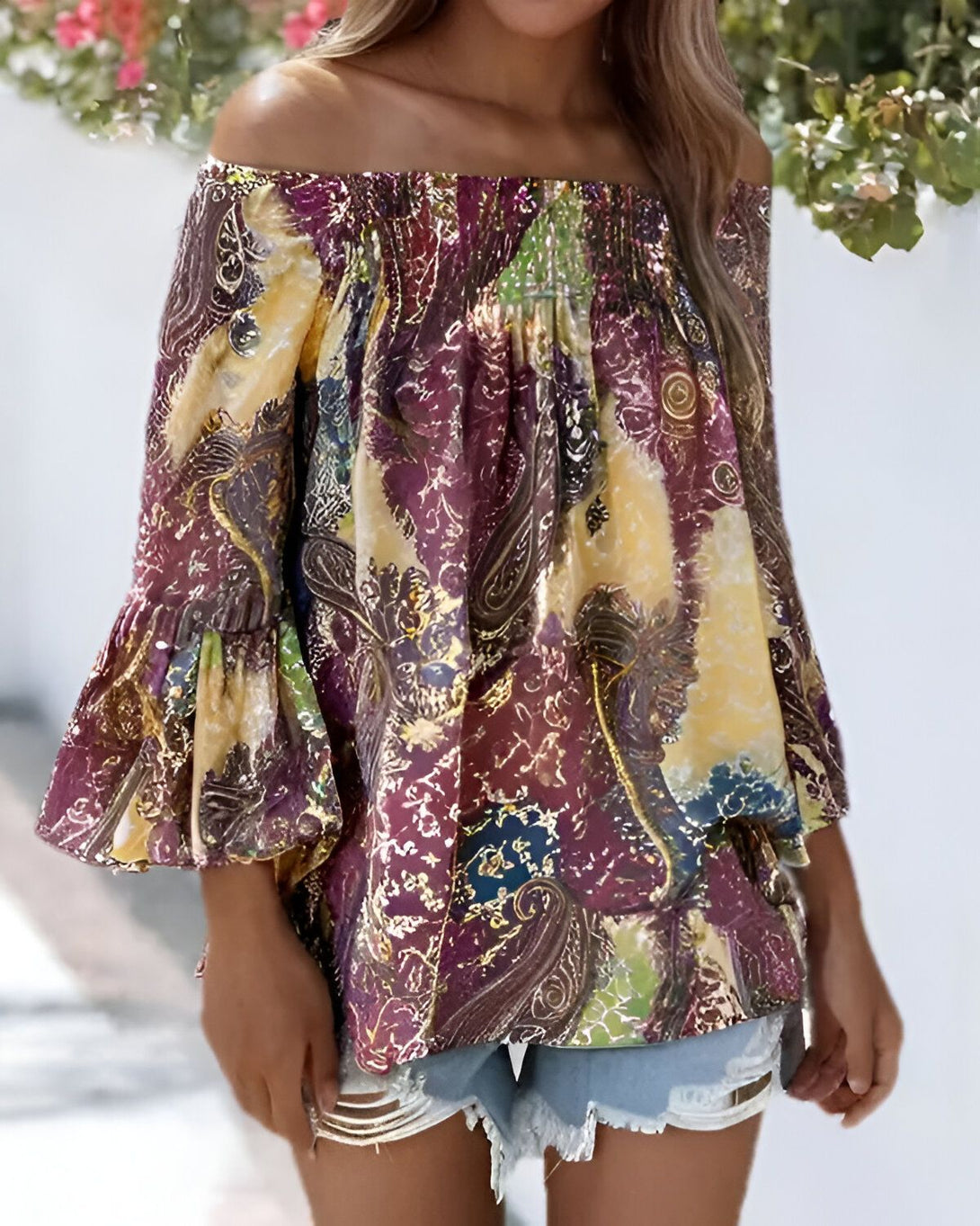 Une femme porte un chemisier jaune et violet à motifs cachemire. Le chemisier est à épaules dénudées et a des manches bouffantes. La femme est également porte un short en jean bleu déchiré.
