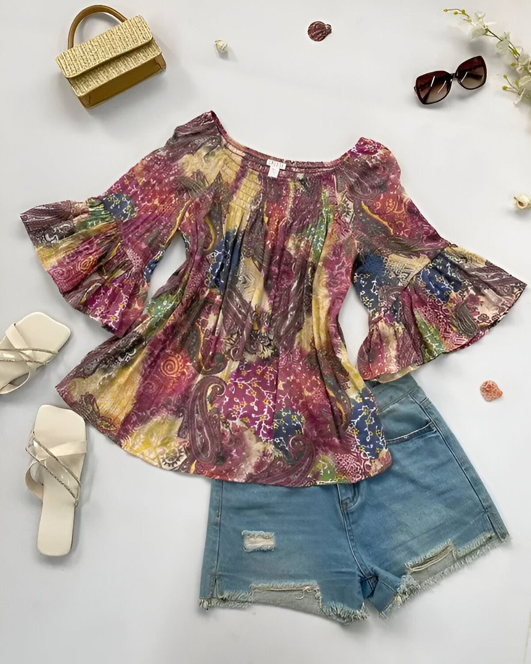 Un chemisier à motifs multicolores avec des manches évasées et un short en jean bleu déchiré. À côté du chemisier, il y a une paire de sandales blanches et des lunettes de soleil marron. En haut à droite, on peut voir un sac à main en paille doré.