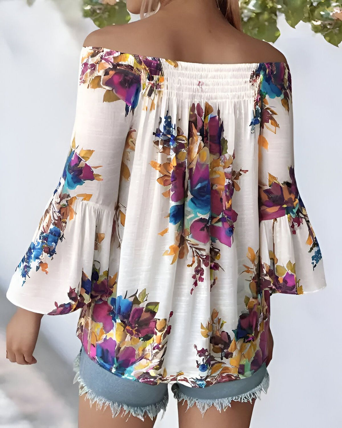 Une femme porte un chemisier à fleurs blanc avec des manches évasées. Le chemisier est décolleté et a une taille élastique. La femme est vue de dos.
