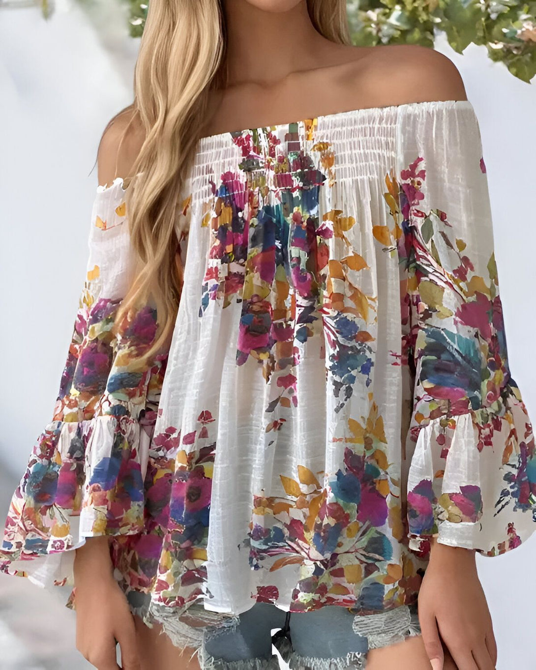 Une jeune femme porte un chemisier à fleurs coloré. Le chemisier est blanc avec des fleurs roses, jaunes, bleues et vertes. Il est à épaules dénudées et a des manches longues et évasées. La femme est blonde et a les cheveux longs et ondulés. Elle porte un short en jean bleu clair et des sandales marron. Elle est debout dans un champ de fleurs et sourit à la caméra.