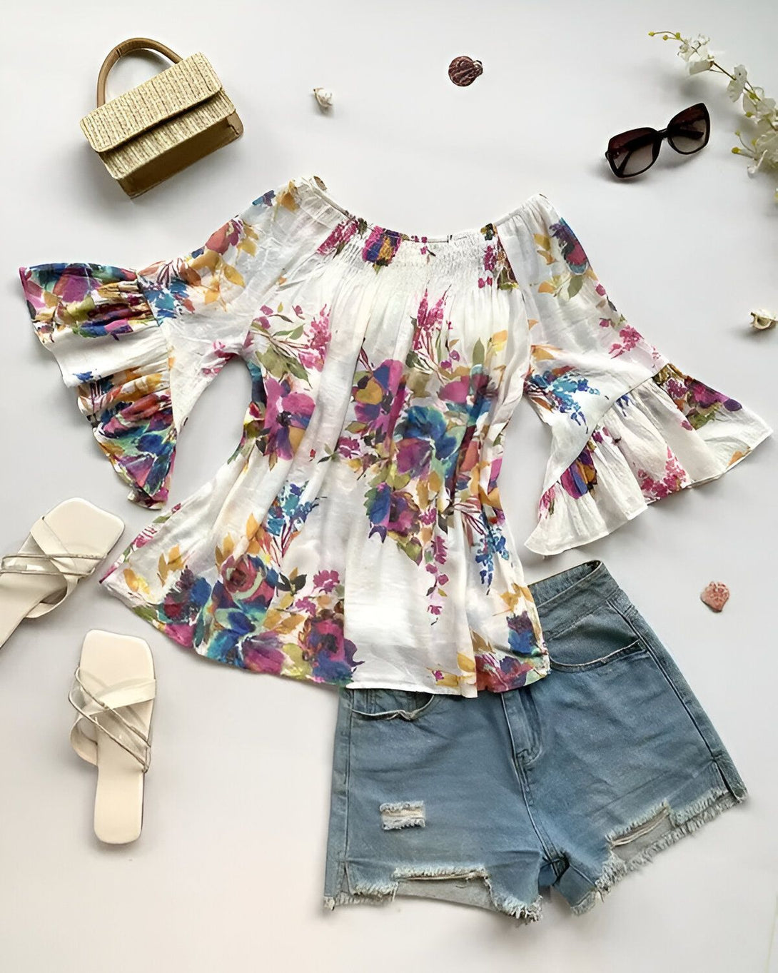 Une image d'un chemisier à fleurs blanc avec des manches bouffantes et un short en jean bleu déchiré. Le chemisier est associé à des sandales blanches et un sac à main en paille beige. Des lunettes de soleil marron et des coquillages sont posés à côté.