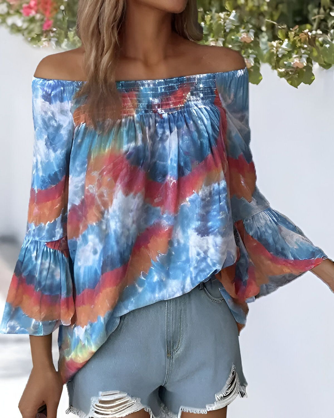 Une jeune femme porte un chemisier tie-dye aux épaules dénudées et des manches évasées. Le chemisier est bleu, orange et blanc. Elle porte également un short en jean bleu déchiré.