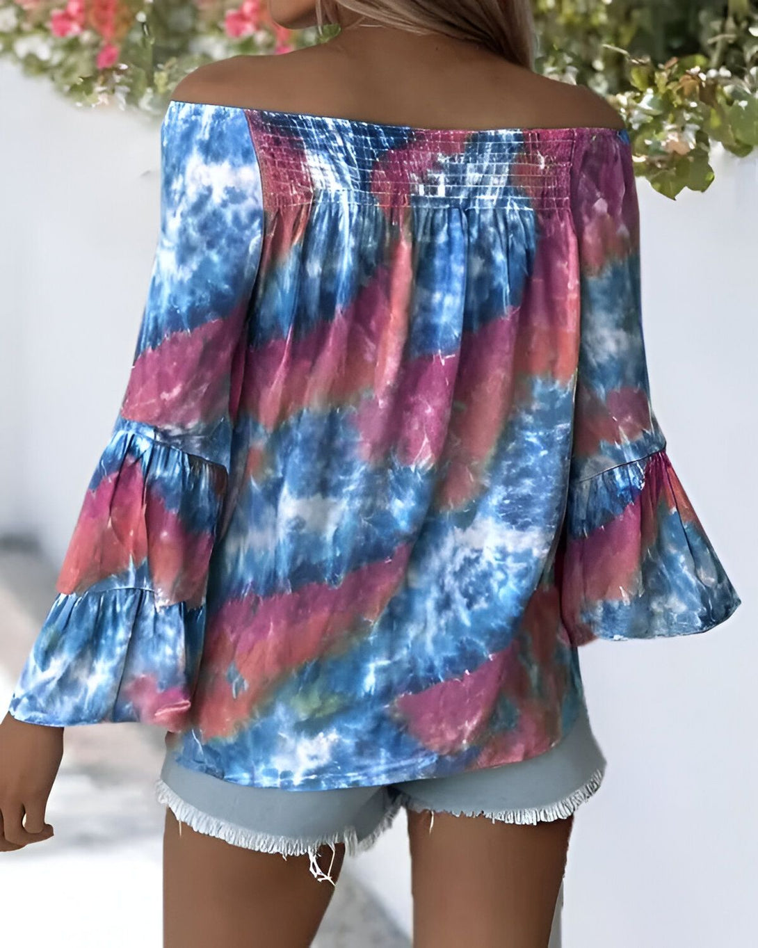 Une femme porte un chemisier tie-dye bleu et rose à épaules dénudées. Le chemisier est ample et a des manches bouffantes. Elle porte également un short en jean bleu clair.