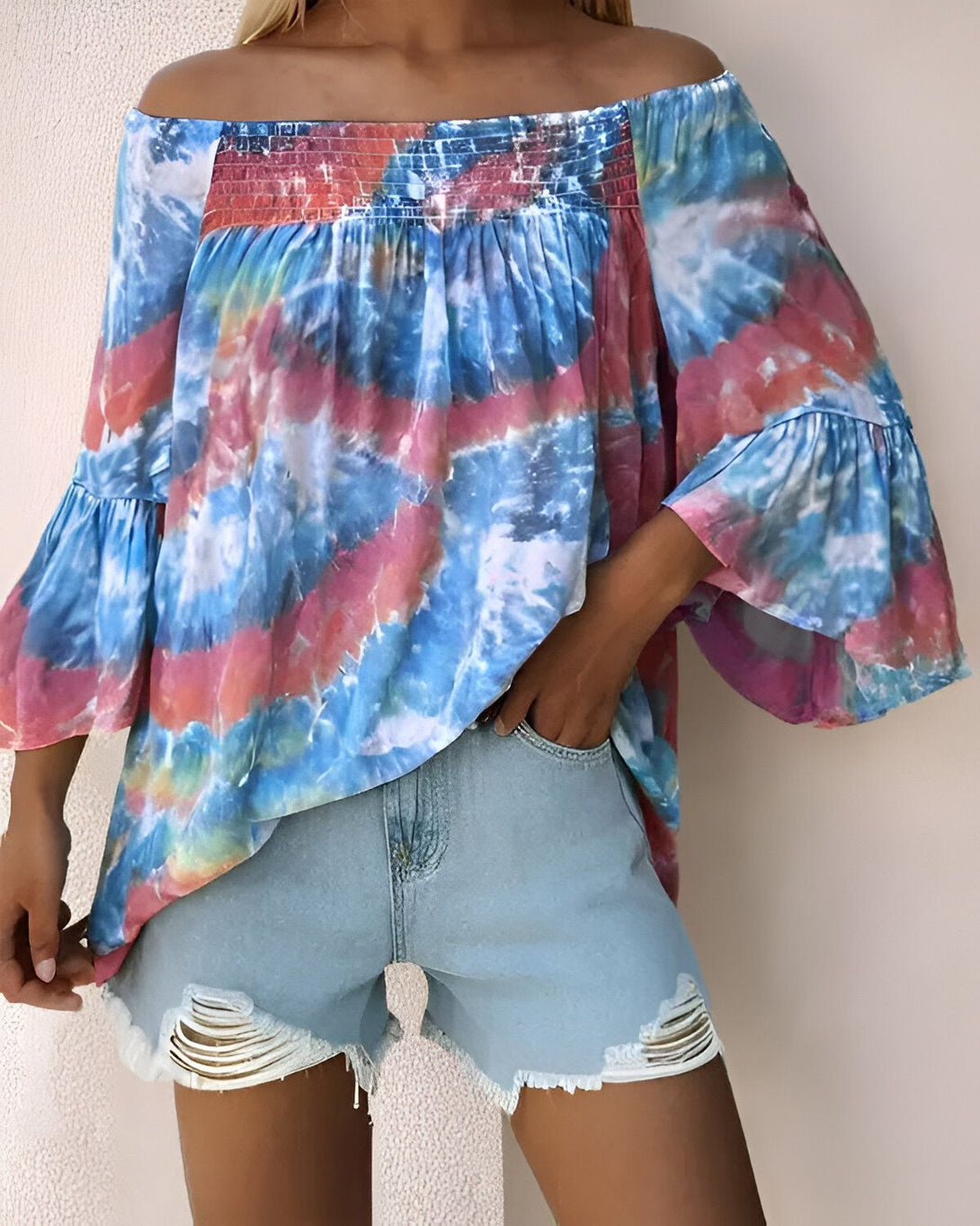 Une femme porte un chemisier tie-dye bleu et rose à épaules dénudées et des shorts en jean déchirés.