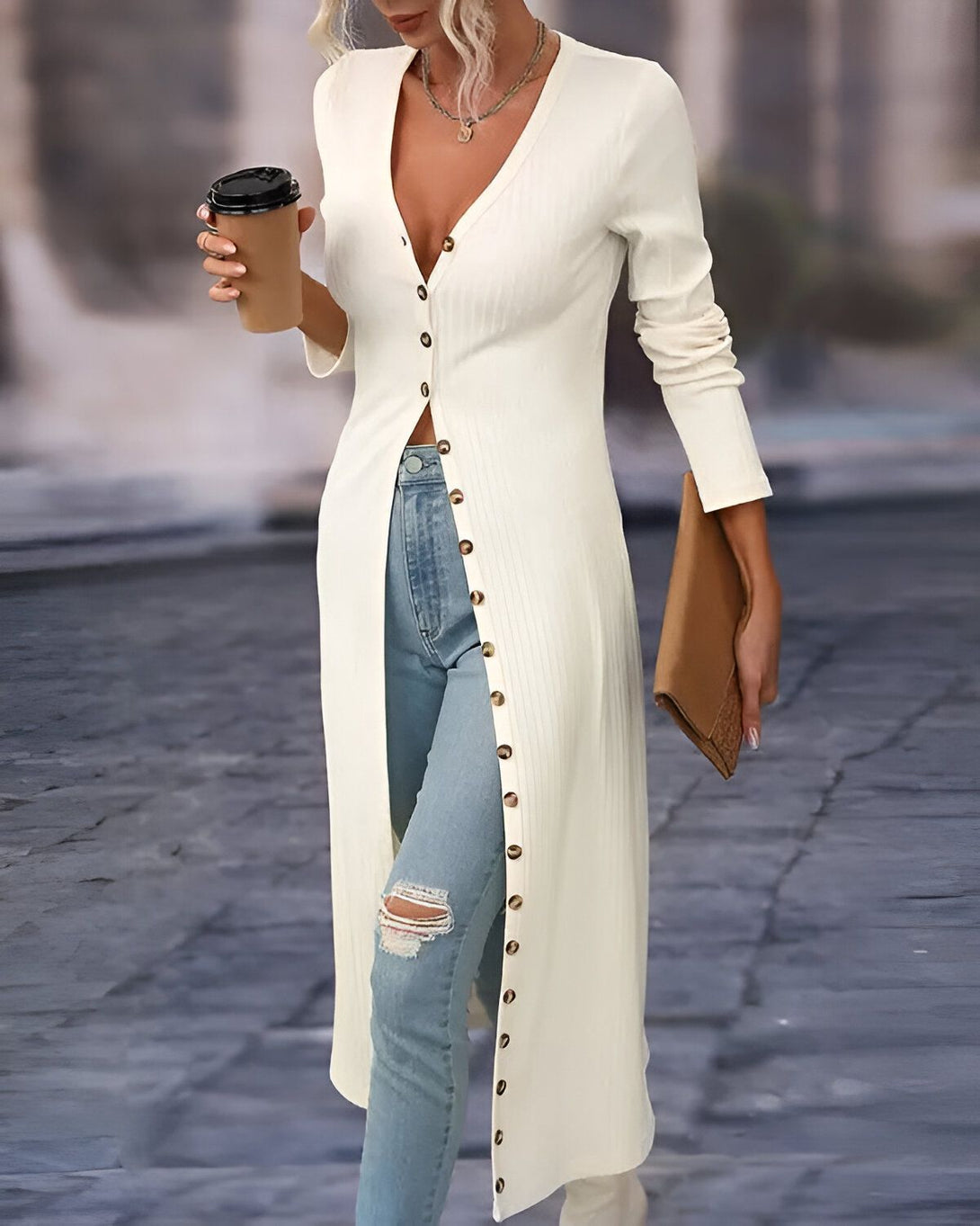 Une femme porte un long cardigan blanc avec des boutons sur le devant. Elle porte également un jean bleu déchiré et des bottes marron. Elle tient une tasse de café à la main.