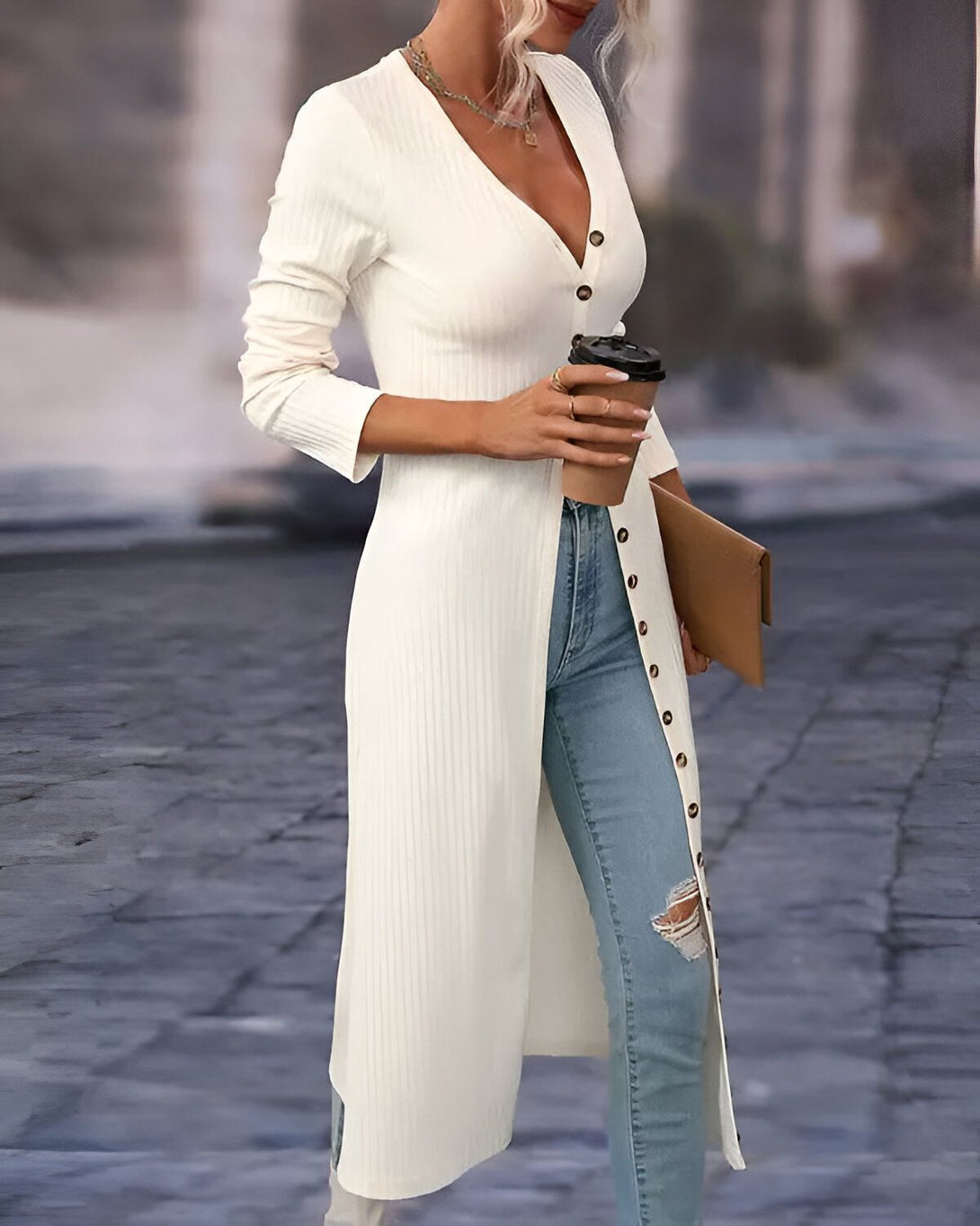 Une femme porte un long gilet blanc avec des boutons sur le devant. Elle porte également un jean bleu déchiré et des talons hauts. Elle tient une tasse de café à la main.