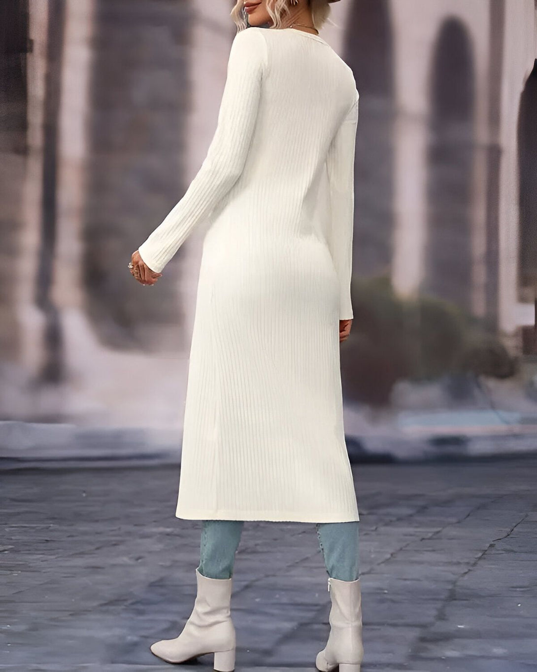 Une femme porte un long gilet blanc en laine côtelée. Il est ouvert sur le devant et possède des manches longues. Elle porte également un jean bleu et des bottes marron clair.