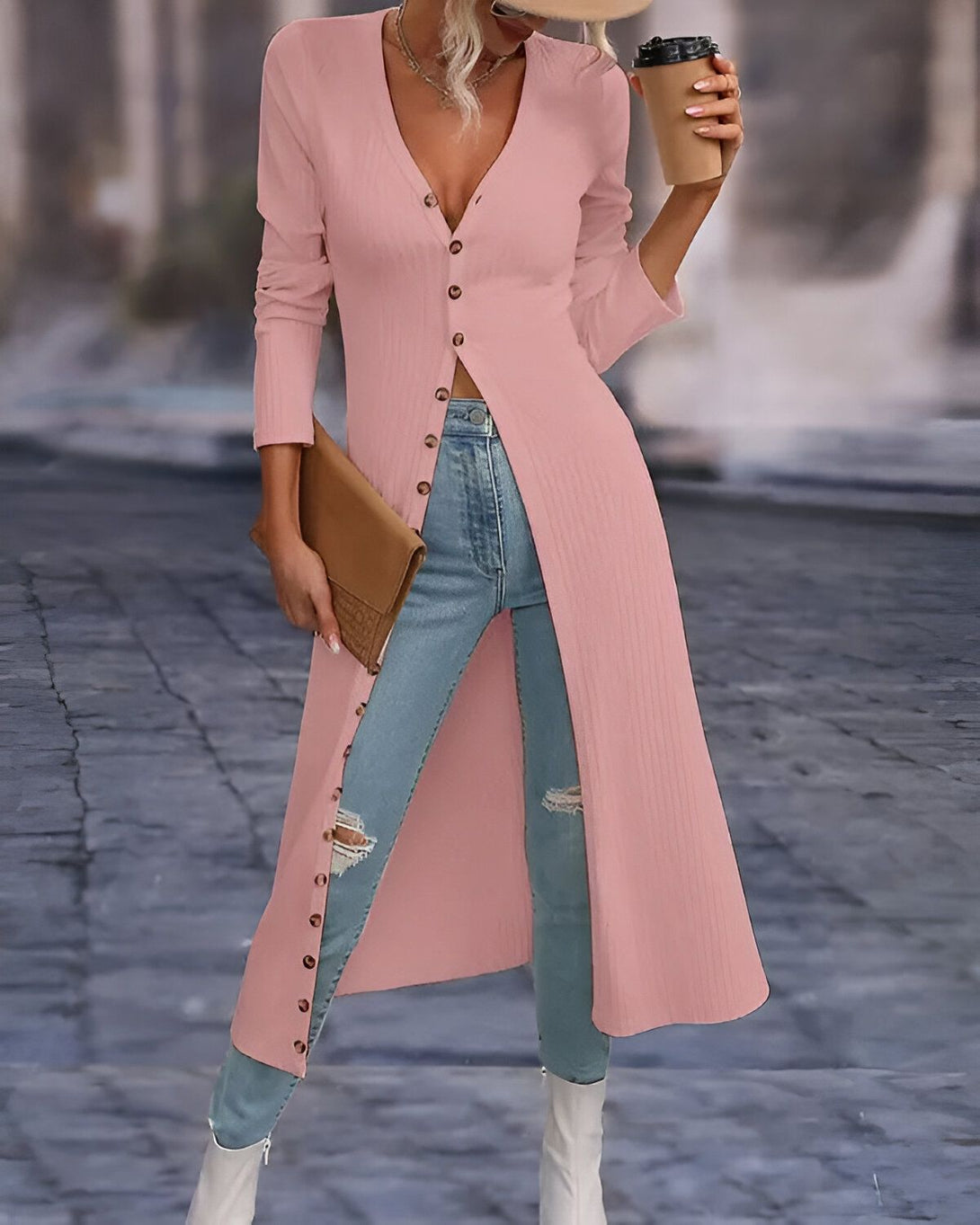 Une femme porte un long cardigan rose boutonné sur un jean bleu déchiré et des bottes blanches. Elle porte un chapeau marron et tient une tasse de café à la main.