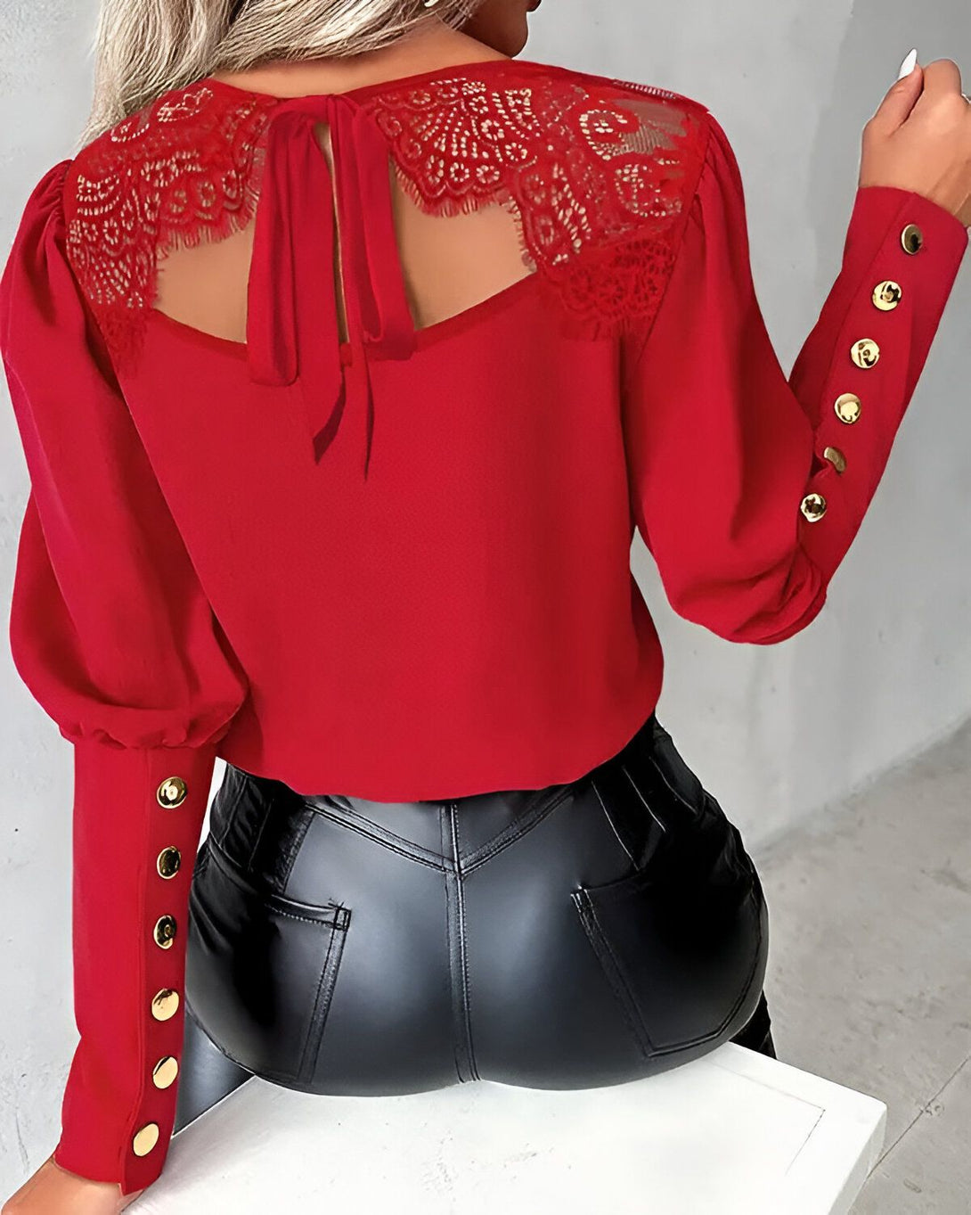Une femme porte un chemisier rouge avec des boutons dorés et un pantalon en cuir noir. Le chemisier est en dentelle et a un nœud dans le dos.