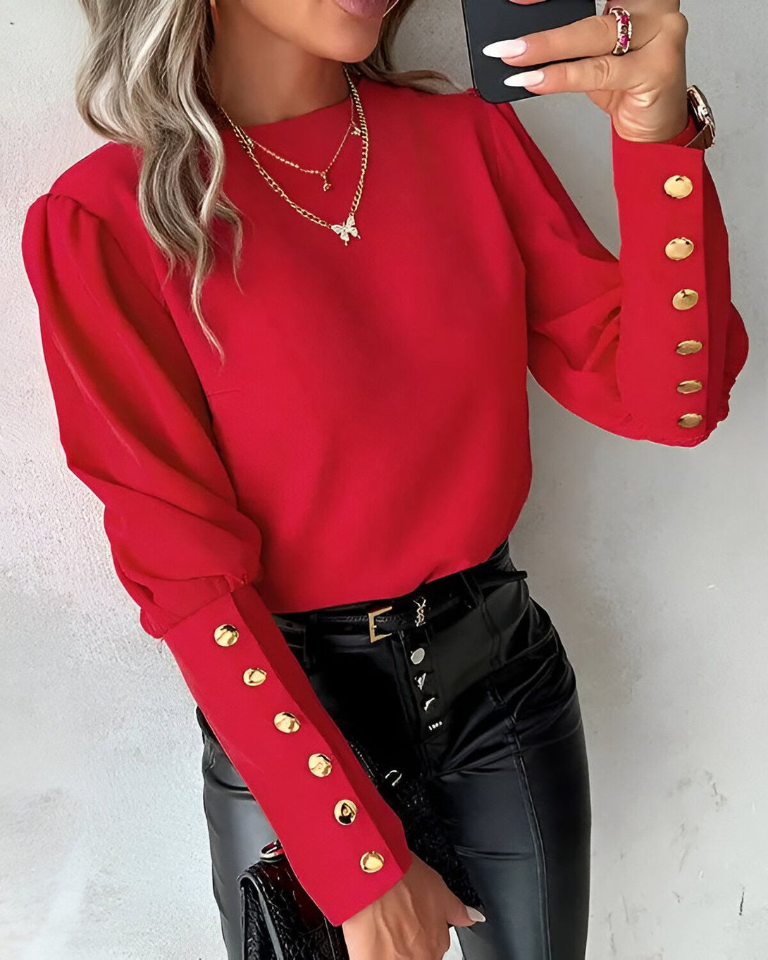 Une femme porte un chemisier rouge à manches bouffantes et boutons dorés. Elle porte également un pantalon en cuir noir et un collier avec un pendentif papillon.