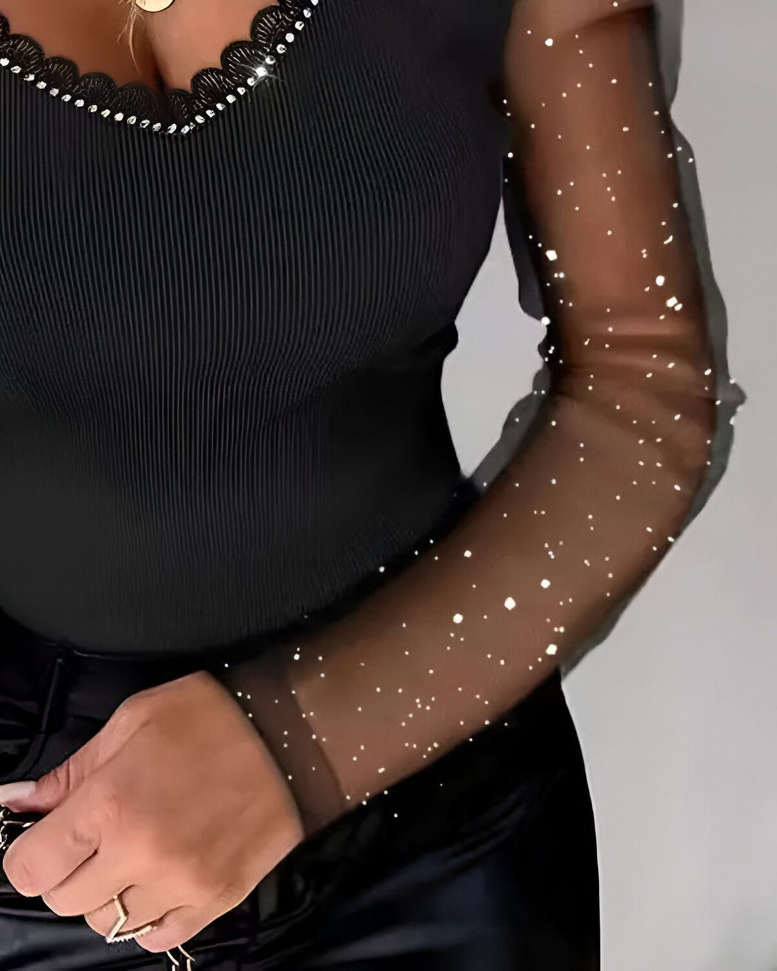 Une femme porte un haut noir à manches longues en résille scintillante. Le haut est ajusté et a un décolleté en V. La femme porte également un pantalon noir.