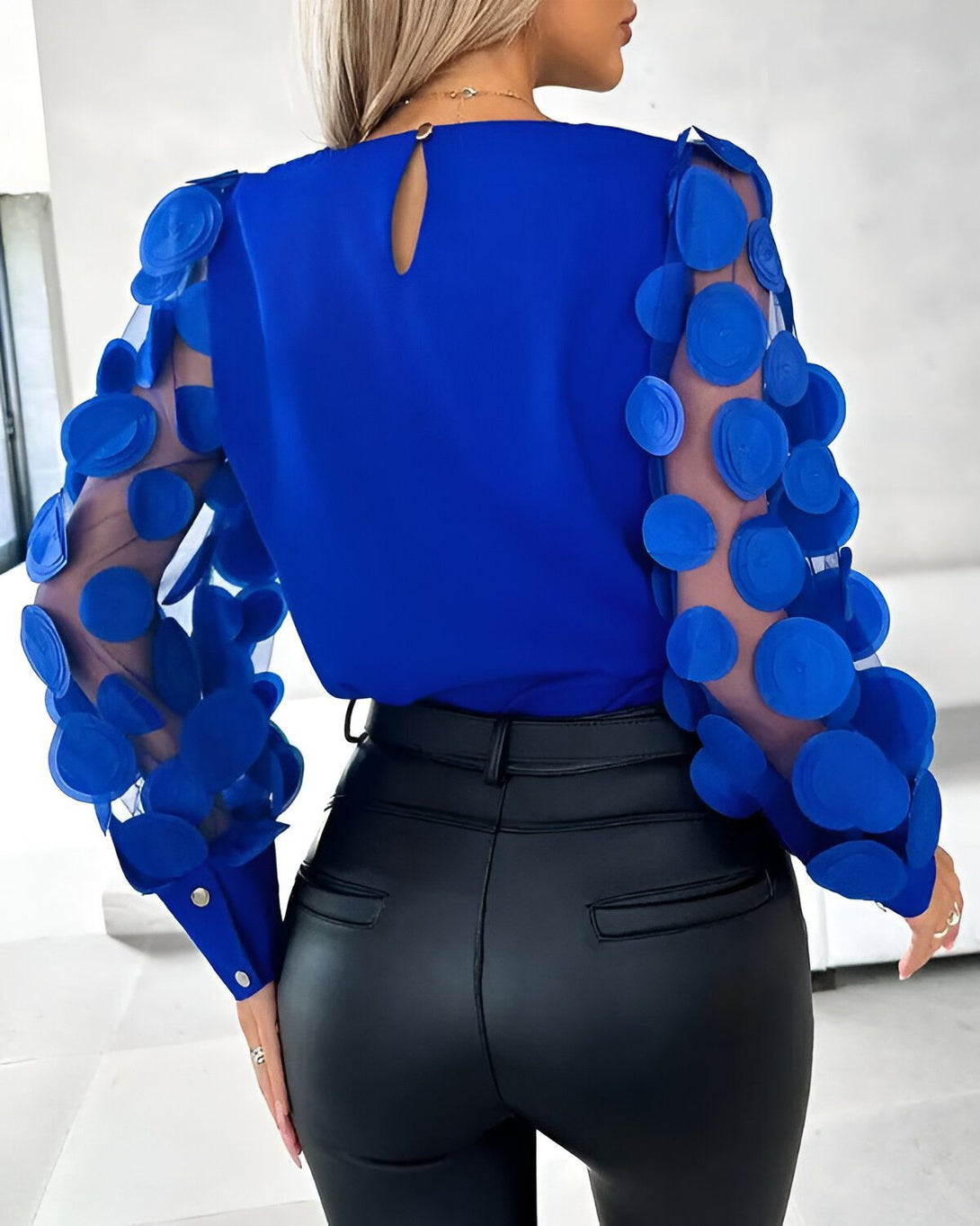 Une femme porte un chemisier bleu avec des manches bouffantes en résille à pois bleus. Le chemisier est rentré dans un pantalon en cuir noir.