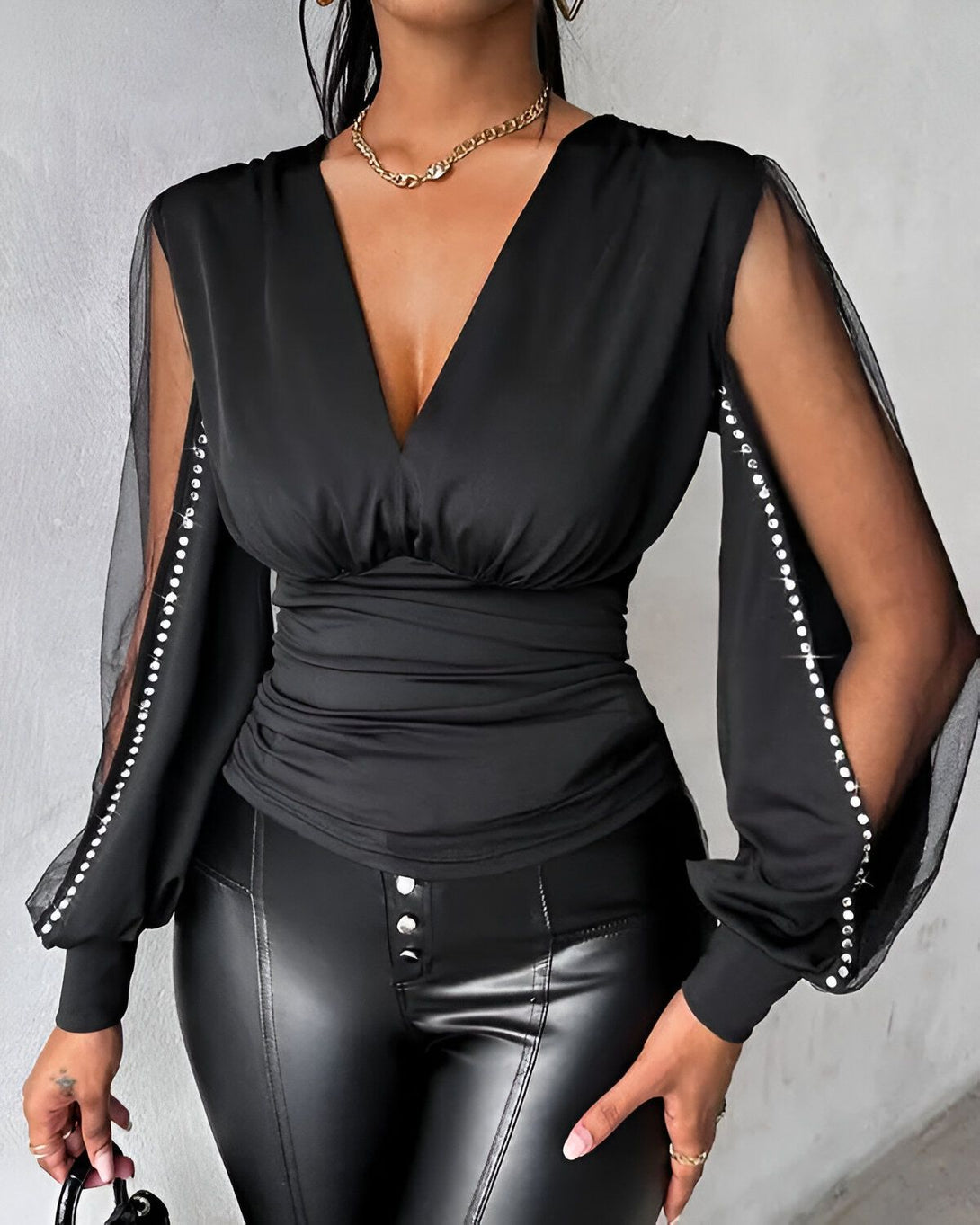 Une femme porte un chemisier noir avec des manches en résille et des strass. Le chemisier est cintré à la taille et a un décolleté en V. Elle porte également un pantalon en cuir noir et un collier doré.