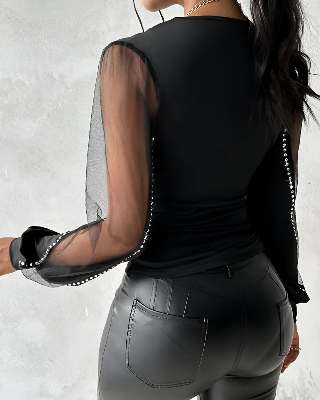 Une femme porte un haut noir moulant avec des manches bouffantes en résille transparente. Le haut est orné de strass sur les manches et dans le dos. Elle porte également un pantalon en cuir noir.
