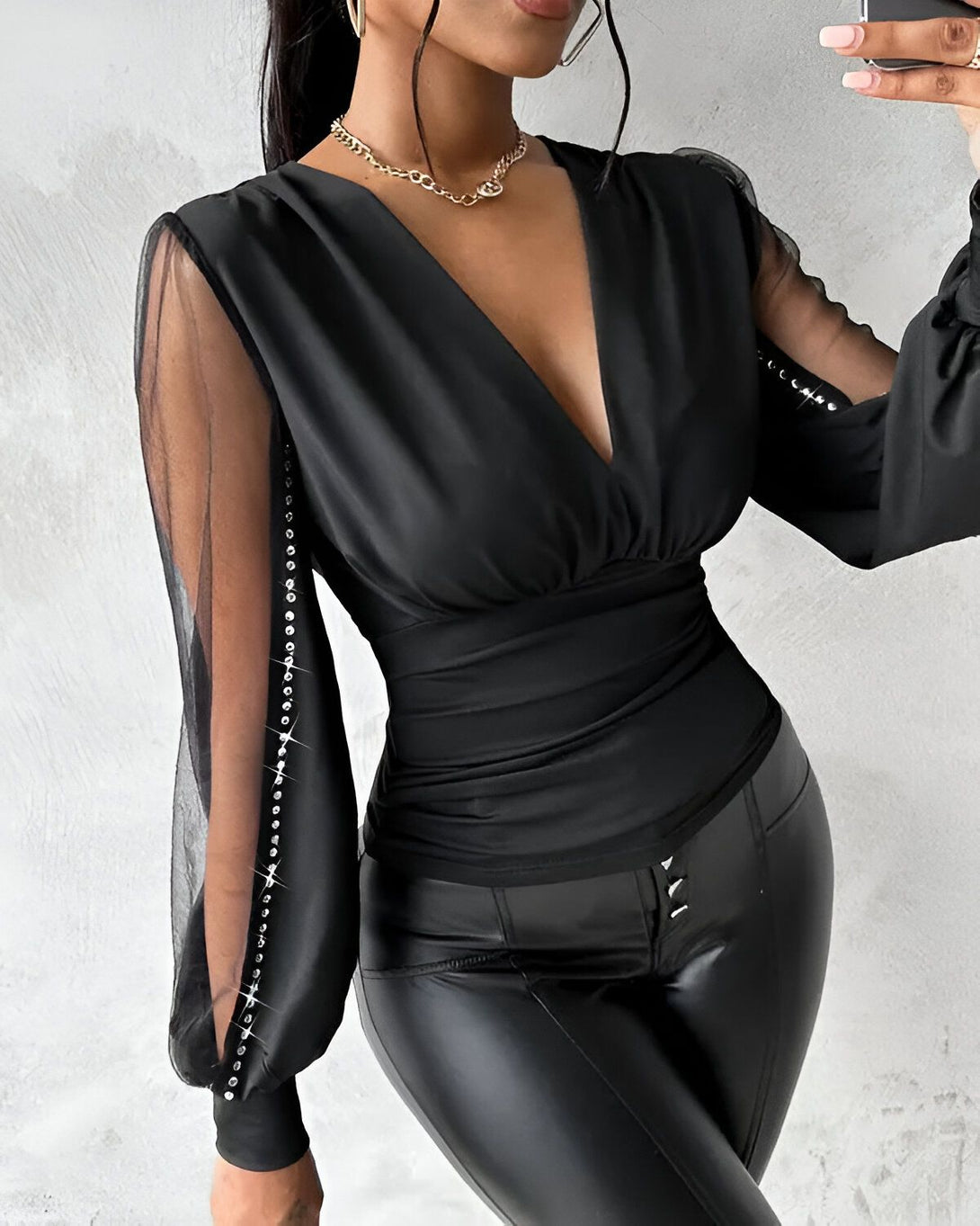 Une femme porte un chemisier noir avec des manches en résille et des boutons en strass. Elle porte également un pantalon en cuir noir.