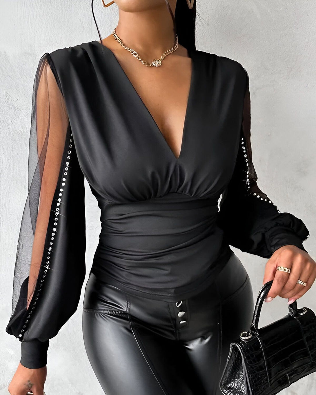 Une femme porte un chemisier noir décolleté en V avec des manches longues en résille et des strass. Elle porte également un pantalon en cuir noir et un collier doré.