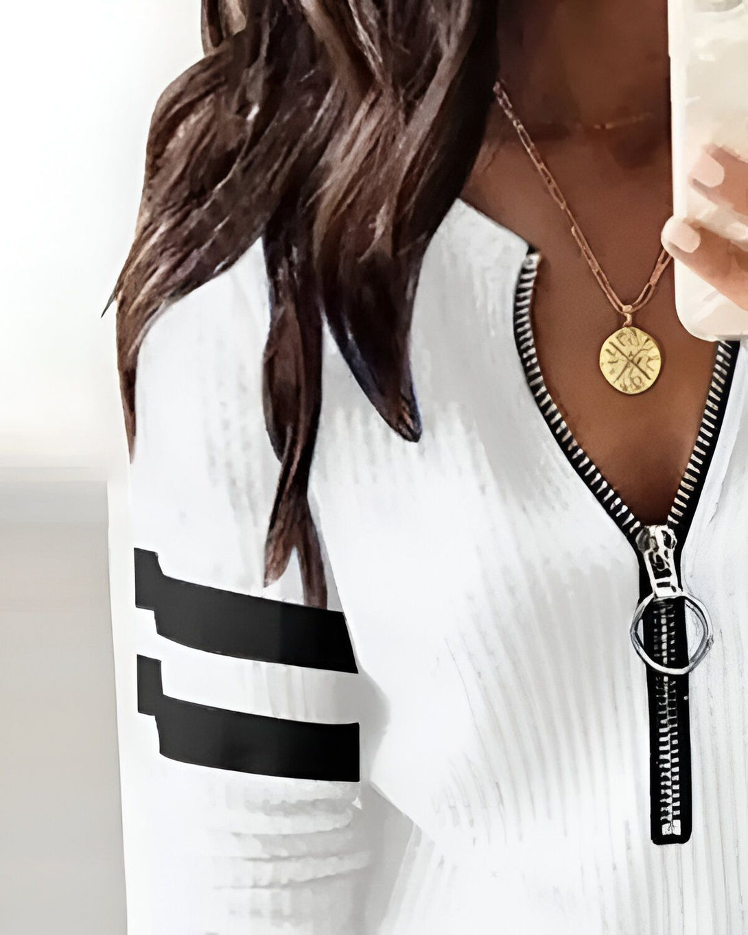 Une femme porte un haut blanc avec des rayures noires sur les manches et un pantalon noir. Elle a les cheveux longs et bruns et porte un collier avec un pendentif rond.