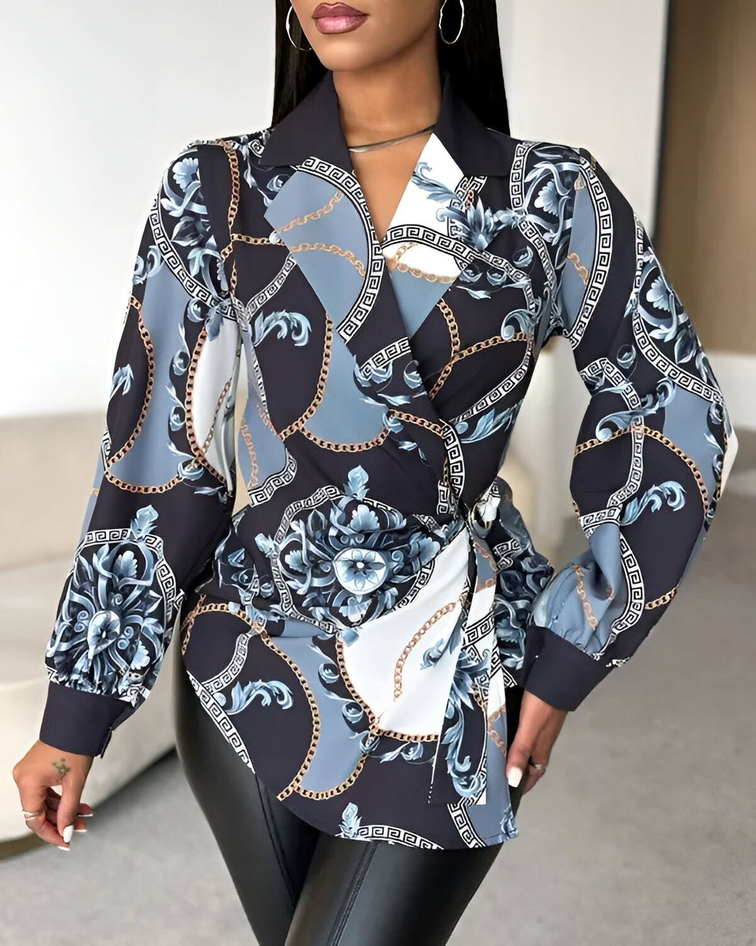 Une femme porte un chemisier noir et blanc à motifs baroques et chaînes dorées. Le chemisier est cintré à la taille et possède un col montant. La femme porte également un pantalon noir en cuir.