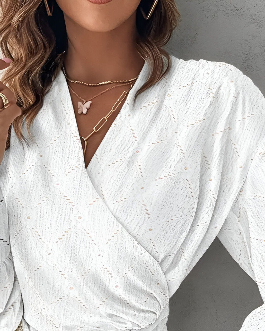 Une femme porte un chemisier blanc à manches longues et un pantalon beige. Elle porte également un collier avec un pendentif en forme de papillon.