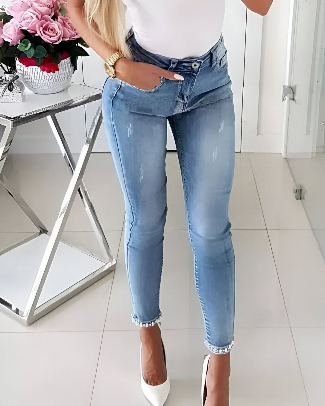Une femme porte un jean bleu moulant avec des perles en bas. Elle porte également un chemisier blanc et des escarpins blancs. Elle est debout dans une pièce avec un sol en marbre blanc et un mur blanc. Il y a une table avec un vase de fleurs roses sur la gauche.