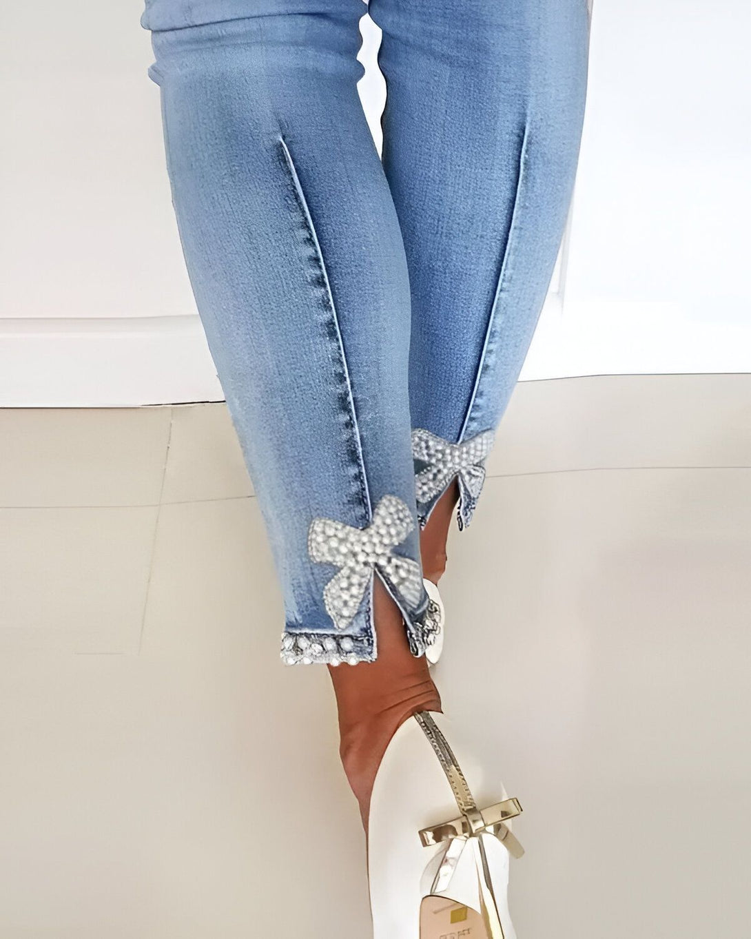 Une femme porte un jean bleu avec des perles et des nœuds sur le bas des jambes. Elle porte des chaussures blanches à talons avec un nœud sur le bout.