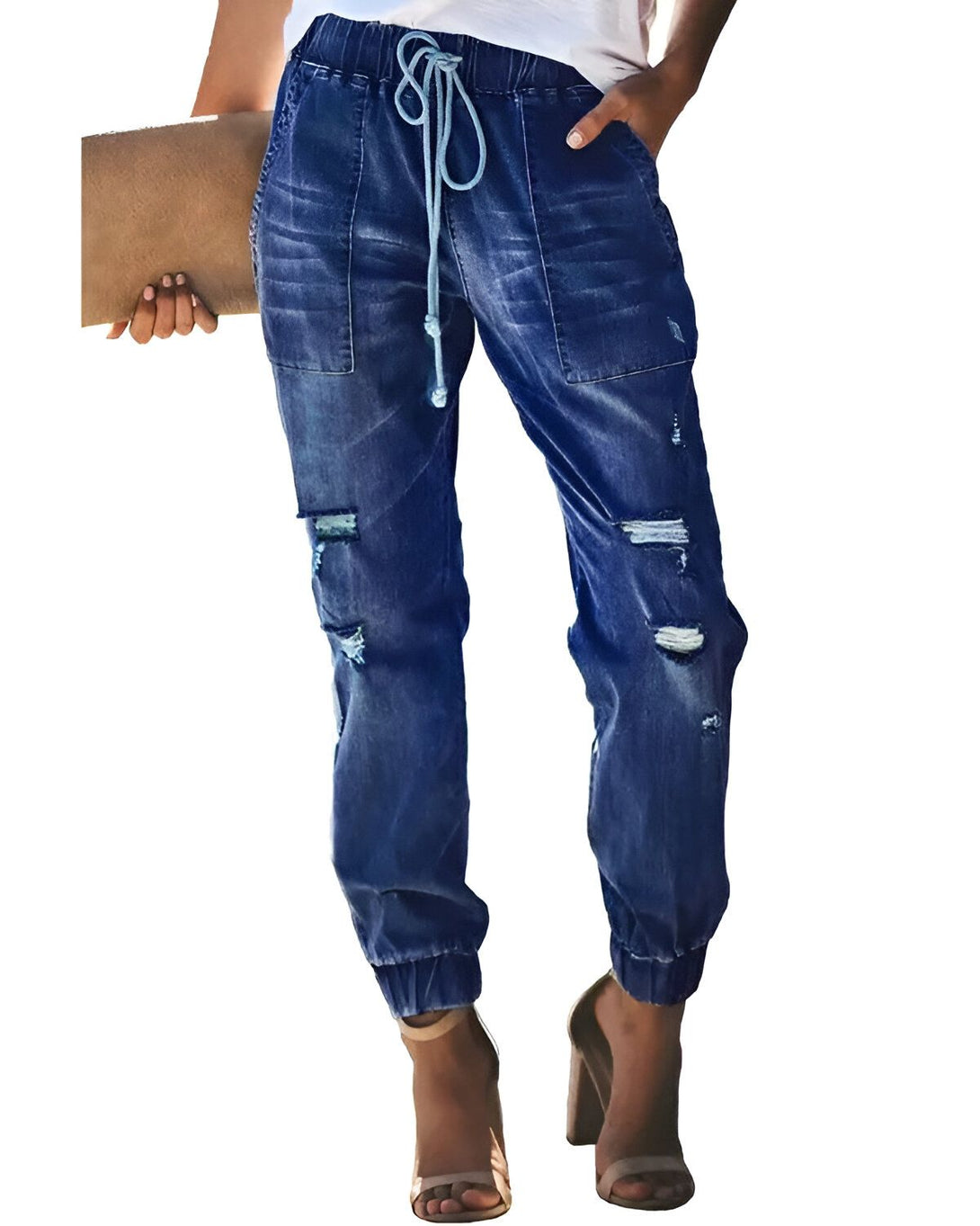 Une femme porte un jean bleu troué et déchiré avec un cordon de serrage à la taille et des talons hauts beiges. Elle porte également un haut blanc et tient un sac à main marron.