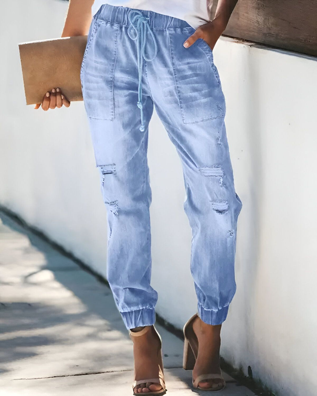 Une femme porte un jean bleu clair déchiré et troué avec un cordon de serrage à la taille et des talons hauts beiges. Elle porte également un haut blanc et tient un livre marron à la main.