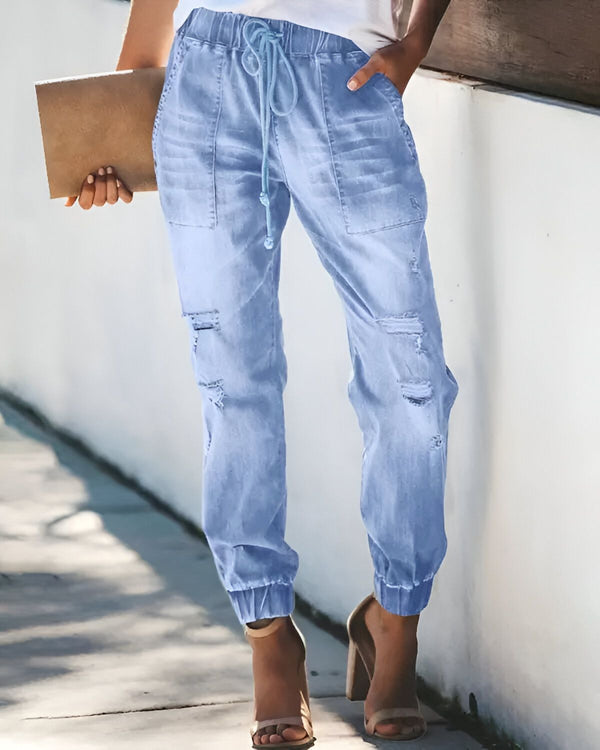 Une femme porte un jean bleu clair déchiré et troué avec un cordon de serrage à la taille et des talons hauts beiges. Elle porte également un haut blanc et tient un livre marron à la main.